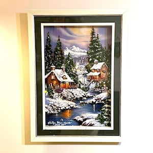 May include: A framed winter landscape print featuring snow-covered cabins, evergreen trees, and a flowing river. The artwork is set against a backdrop of snow-capped mountains and a colorful sky. The frame is white.