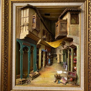 May include: A framed 3D relief sculpture of a Middle Eastern marketplace. The artwork shows detailed buildings, balconies, and shops with a warm colour palette of browns, greens, and yellows. The frame is ornate with a gold finish.