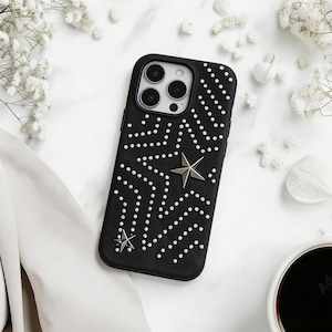 May include: Black phone case featuring a dotted design and two silver star decorations. The case is surrounded by white flowers and a cup of coffee. The case is a stylish accessory.