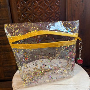 May include: A clear, rectangular vinyl pouch featuring a yellow zipper closure and a small red apple charm attached to the zipper pull. The pouch is filled with colorful, scattered confetti in the shapes of letters, pencils, and glittery gold dots.