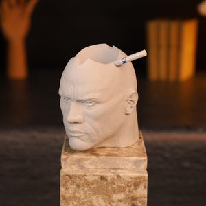 Può includere: Una testa scolpita di colore chiaro con una parte superiore scavata, che assomiglia a un attore famoso. Una sigaretta poggia nell'apertura della testa. La scultura poggia su una base quadrata strutturata, suggerendo un oggetto decorativo o un posacenere unico.