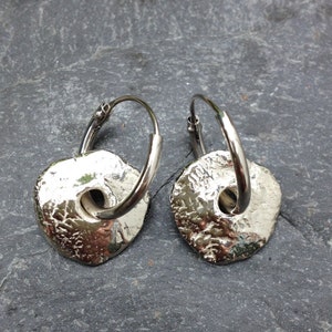 May include: A pair of silver hoop earrings with textured, circular, silver charms. The charms have a hole in the center.