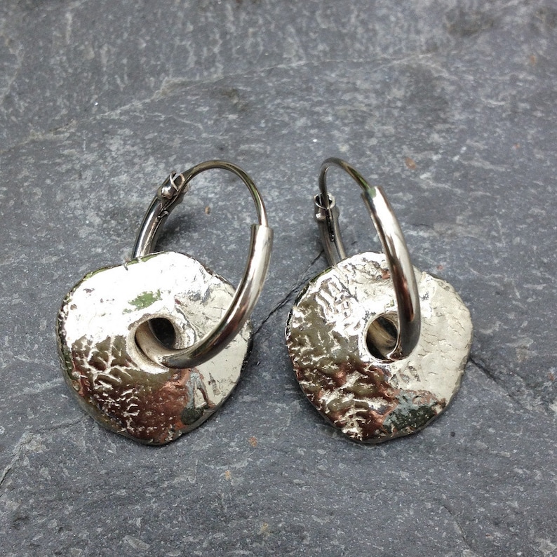 May include: A pair of silver hoop earrings with textured, circular, silver charms. The charms have a hole in the center.