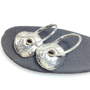 May include: A pair of silver hoop earrings with textured, circular pendants. The earrings are displayed on a dark gray stone surface.