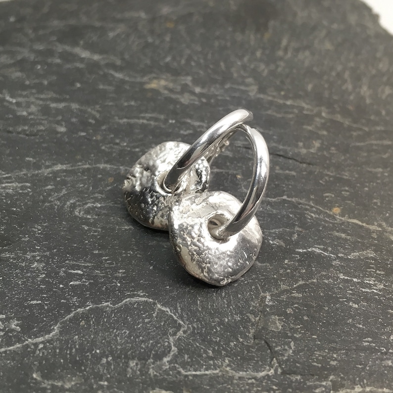 May include: A pair of silver hoop earrings with textured, round, disc-shaped charms.