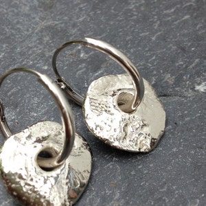 May include: A pair of silver hoop earrings with textured, round, silver charms. The charms have a hole in the center.