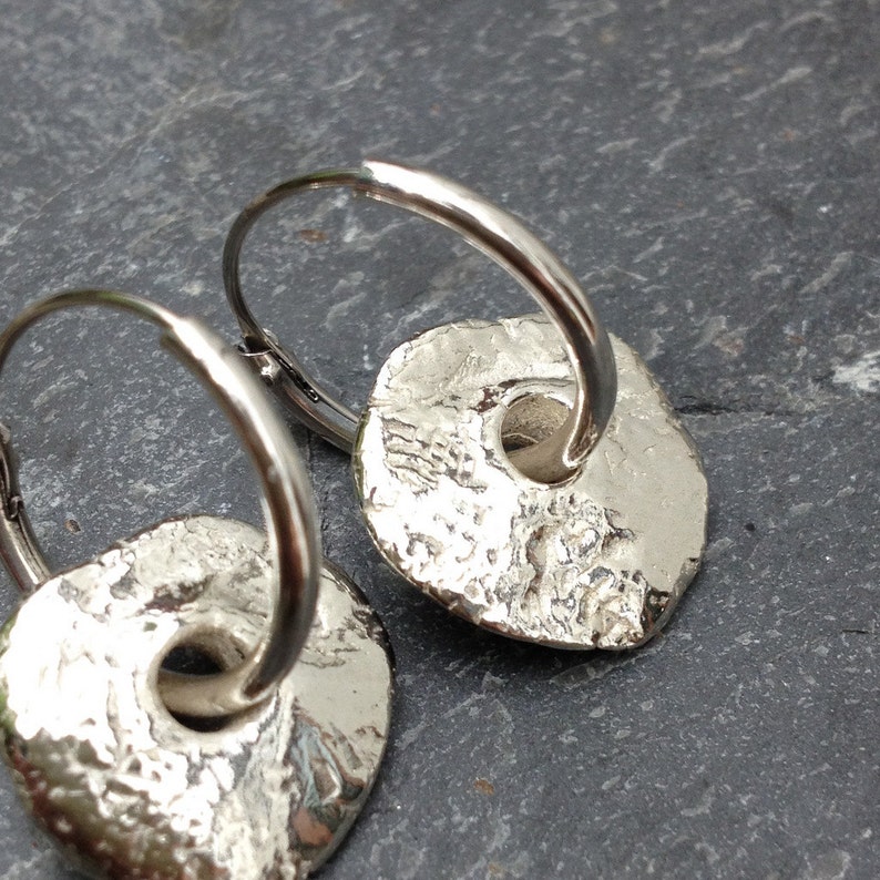 May include: A pair of silver hoop earrings with textured, round, silver charms. The charms have a hole in the center.