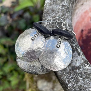 May include: Silver hammered disc earrings with three small rivets and a black stone accent. The earrings have a textured surface and are suspended from a silver hook.