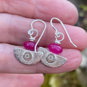 May include: A pair of silver earrings with a half-moon design. Each earring features a textured silver crescent shape with a central circular accent and a vibrant magenta bead. The earrings have a hook closure.