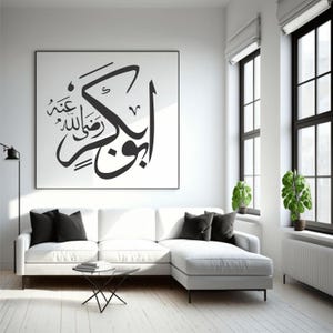 May include: A modern living room featuring a white sectional sofa with black throw pillows, a small glass table, and a large framed artwork with Arabic calligraphy in black. The room has white walls, black window frames, and potted plants.