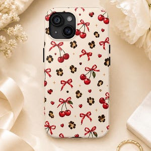 Puede incluir: Una funda de smartphone de color crema con un patrón repetitivo de cerezas rojas, lazos rojos, pequeñas manchas de estampado de leopardo y diminutos corazones rojos. El texto THE X K STUDIO está impreso en letras negras pequeñas en la parte inferior.