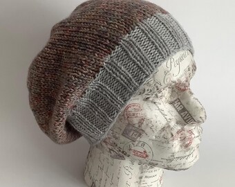 May include: A handmade, knitted beret in shades of brown, gray, and red. The beret features a ribbed gray brim and a textured, slouchy crown. The hat is displayed on a decorative head form.