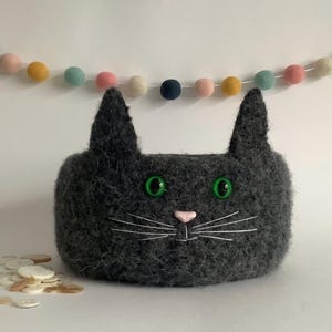 May include: A dark grey cat-shaped container with green eyes and a pink nose. White whiskers are stitched on the face. A colourful garland of felt balls hangs in the background. A pile of buttons is in the foreground.