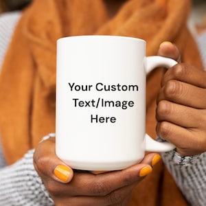 May include: A white ceramic mug with a large handle, featuring the text "Your Custom Text/Image Here" in black. The mug is held by a person with orange nail polish, against a blurred background.