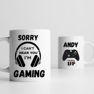 Peut inclure: Deux mugs en céramique blancs. L'un porte l'inscription "SORRY I CAN'T HEAR YOU I'M GAMING" avec un motif de casque. L'autre mug porte le nom "ANDY" et un motif de manette de jeu vidéo avec "LEVEL UP".