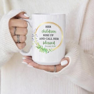 May include: White ceramic mug featuring the text "HER children RISE UP and CALL HER blessed" in green and gold, with floral details. The mug is held by a person wearing a white jumper.