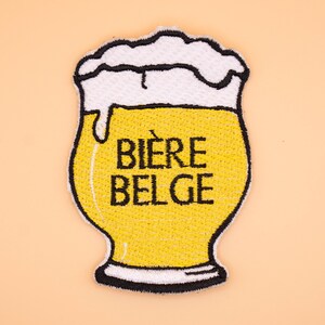Puede incluir: Un parche bordado con forma de vaso de cerveza con una espesa espuma blanca. El líquido es amarillo y el texto BIÈRE BELGE está bordado en negro en el centro. Todo el diseño está delineado con hilo negro sobre un fondo de color claro.
