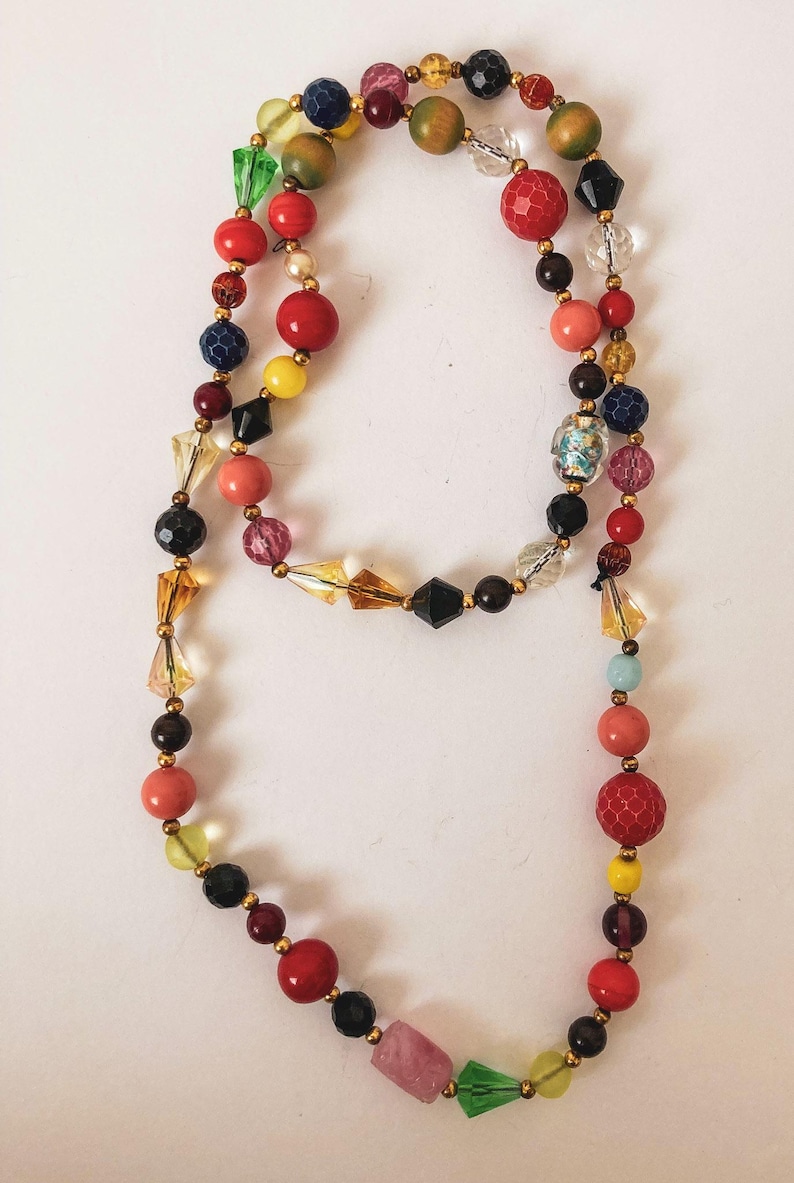Puede incluir: Un collar de cuentas colorido con una variedad de formas y tama&ntilde;os. El collar presenta cuentas rojas, amarillas, azules, verdes y rosas, intercaladas con espaciadores dorados. Las cuentas est&aacute;n dispuestas en un dise&ntilde;o largo y fluido.
