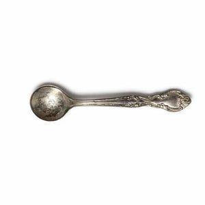 Può includere: Un piccolo cucchiaino d'argento antico con una coppetta arrotondata e un manico ornato. Il cucchiaino è probabilmente in argento sterling e presenta dettagliati motivi floreali e a volute. Il cucchiaino è lungo circa 13 cm.