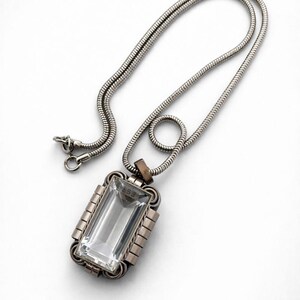 Puede incluir: Un collar plateado con un colgante rectangular. El colgante presenta una piedra rectangular grande y transparente engastada en un marco detallado. La cadena tiene un diseño fino y texturizado. El colgante tiene el número "653" grabado.