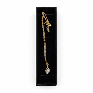 Puede incluir: Un collar dorado con un colgante de piedra preciosa azul claro, presentado en una caja de joyería negra. La cadena es fina y el colgante tiene forma de lágrima. La caja rectangular tiene un interior aterciopelado.