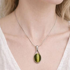Puede incluir: Un collar de plata con un colgante en forma de lágrima con una piedra preciosa verde ojo de gato. El colgante cuelga de una fina cadena de plata. La piedra preciosa tiene una franja vertical de luz.