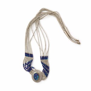 Puede incluir: Collar plateado con múltiples hilos de finas cadenas de cuentas. El collar tiene un colgante central ovalado con una piedra azul y está adornado con cuentas azules. El diseño es intrincado y detallado.