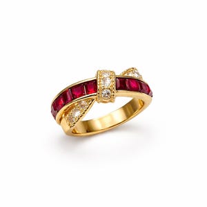Può includere: Un anello d'oro con un design a fiocco, con pietre rosse quadrate e piccole pietre bianche. L'anello ha una finitura lucida ed è posto su uno sfondo bianco.