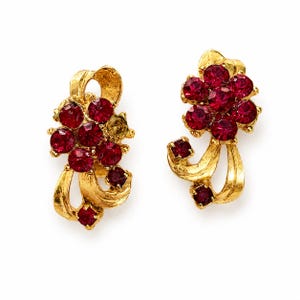 May include: A pair of gold-toned clip-on earrings. Each earring features a cluster of round, deep red gemstones and a few square-cut gems. The design includes a decorative gold ribbon element, creating a vintage aesthetic.