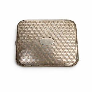 May include: A silver rectangular cigarette case with a geometric pattern on the front and a small oval emblem. The case has a decorative border and a hinged closure. The case is likely made of metal.