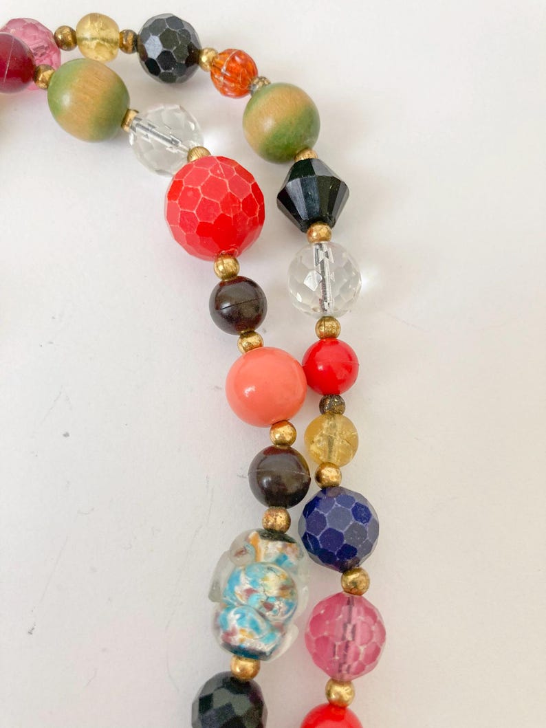 Puede incluir: Un collar de cuentas de colores con una variedad de formas y tama&ntilde;os. Las cuentas son en tonos de rojo, rosa, naranja, verde, negro, azul y transparente. Espaciadores dorados separan las cuentas. El collar est&aacute; sobre un fondo blanco.