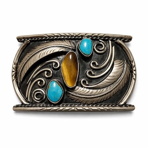 May include: A silver-toned belt buckle with an oval shape, featuring a central brown stone and two turquoise stones. The design includes feather and swirl details, with a textured border.