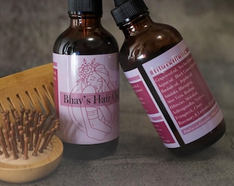 May include: Two amber glass dropper bottles labeled Bhav's Hair Oil with pink labels featuring a line art illustration of a person and a list of ingredients including Grapeseed, Argan, and Jojoba oils, positioned next to a round bamboo hair brush.