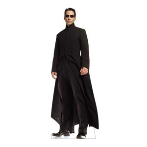 May include: A life-sized cardboard cutout of a person with short dark hair, wearing dark sunglasses, a long black high-collar coat, matching black trousers, and black dress shoes. The figure stands in a neutral pose against a plain white background.