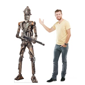 May include: A life-size cardboard cutout of the IG-11 droid from The Mandalorian. The droid is metallic with a gun and a bandolier. A person is standing next to the cutout, gesturing towards it.