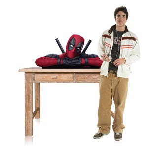 May include: A Deadpool bust figure featuring the character in a red and black suit with crossed swords on the back, resting its arms on a wooden table. The figure is displayed alongside a person wearing a white jacket, black shirt, and tan trousers.