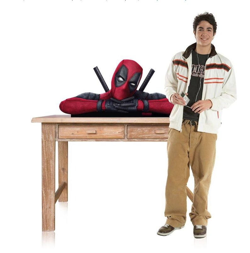 May include: A Deadpool bust figure featuring the character in a red and black suit with crossed swords on the back, resting its arms on a wooden table. The figure is displayed alongside a person wearing a white jacket, black shirt, and tan trousers.