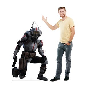 May include: A life-size cutout of a character in a futuristic military suit, kneeling on one knee. The suit is primarily gray with red accents. A man in a yellow shirt and jeans stands next to the cutout, gesturing towards it.