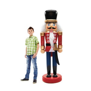 May include: A large, decorative wooden nutcracker soldier in a red and black uniform, standing on a red base. The nutcracker has a white beard and a black hat with gold accents. A young person stands next to the nutcracker.
