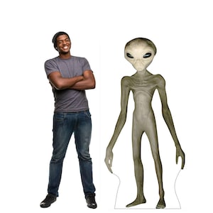 May include: A life-size cutout of a gray alien with large black eyes and a slender physique stands next to a person wearing a gray t-shirt, blue jeans, and a black beanie. The alien has long arms and legs.