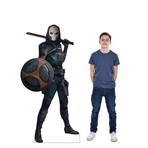 May include: A life-size cardboard cutout of a character in a black and blue costume, holding a sword and shield. The character wears a white mask and hood. A person in a blue t-shirt and jeans stands next to the cutout.