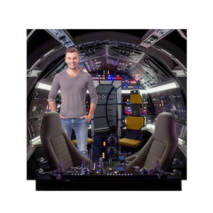 May include: A person stands in a spaceship cockpit with a gray v-neck shirt and blue jeans. The cockpit features control panels, yellow seats, and a futuristic design. The background is dark with various lights.