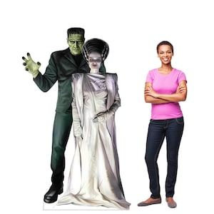 May include: Cardboard cutout of Frankenstein and his bride, with a woman standing next to them. Frankenstein is in a black suit with green skin. The bride wears a white gown. The woman is wearing a pink shirt and blue jeans.