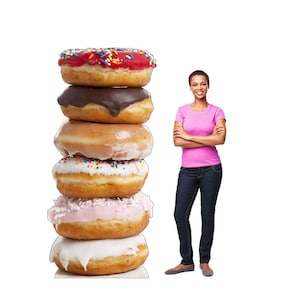 May include: A stack of six donuts with various toppings, including sprinkles, chocolate frosting, and pink icing. A woman in a pink shirt and jeans stands next to the donuts.