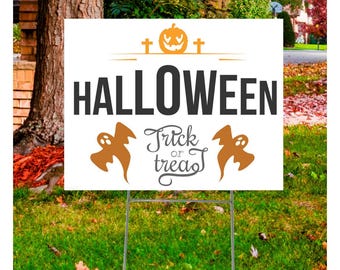 May include: A white Halloween yard sign with a pumpkin and cross design above the word "HALLOWEEN" in black. Below, it says "Trick or Treat" with two orange ghost figures on either side. The sign is on a metal stand in a grassy yard.