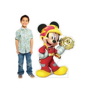 May include: A life-size cardboard cutout of Mickey Mouse in a red racing suit and yellow helmet, holding a gold trophy. The cutout is next to a young boy wearing a floral button-down shirt and blue jeans.