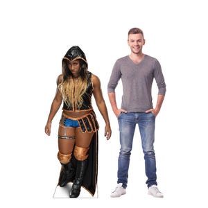 May include: A cardboard cutout of a person in a black and gold costume with a hood, standing next to a man in a gray shirt and jeans. The costume includes a sleeveless top, shorts, and knee-high boots. The man is smiling.