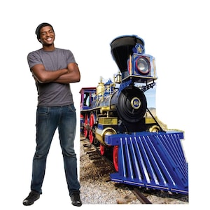 May include: A blue, black, and gold toy train with red wheels. The train has the number 60 on the front. The train is on a track. The train is next to a person wearing a gray shirt, blue jeans, and a black beanie.