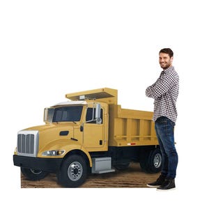 May include: A yellow dump truck with a silver grille and black tyres, positioned next to a life-sized cardboard cutout of a person wearing a plaid button-down shirt and blue jeans, standing with arms crossed on a brown dirt-textured surface.