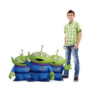 May include: Three green alien figures with three eyes, wearing blue and purple outfits, stand in front of a person wearing a green plaid shirt and blue jeans. The aliens have antennas and are in a waving pose.